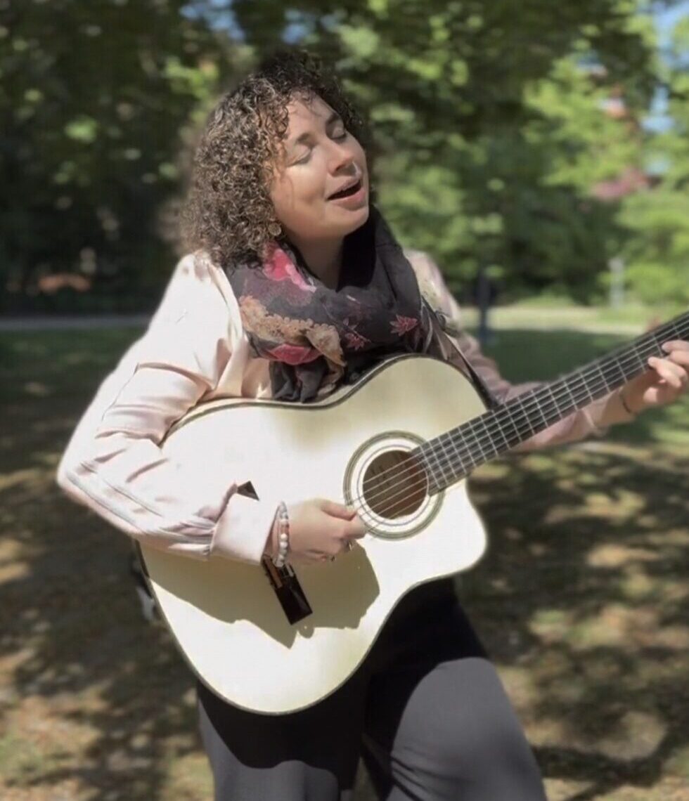 Sängerin ILVA spielt Gitarre und singt im Freien, umgeben von grünen Bäumen an einem sonnigen Tag.