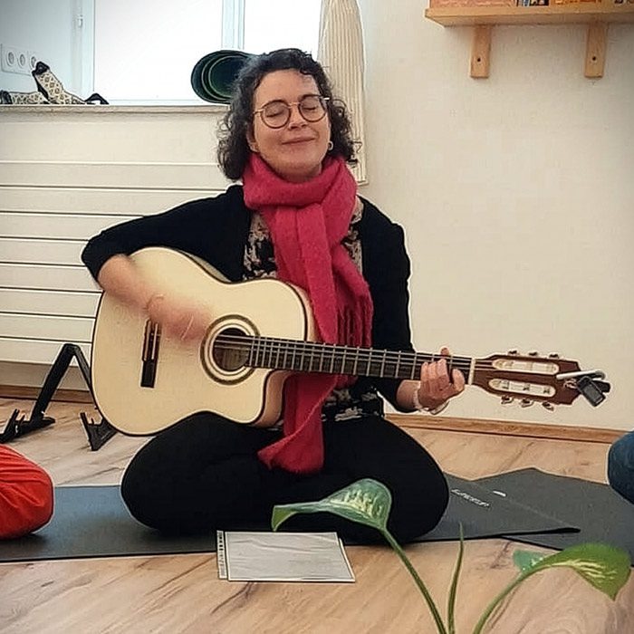 Frau sitzt im Schneidersitz auf einer Matte und spielt Gitarre, während sie mit geschlossenen Augen singt; ruhige, gemütliche Atmosphäre in einem hellen Raum.